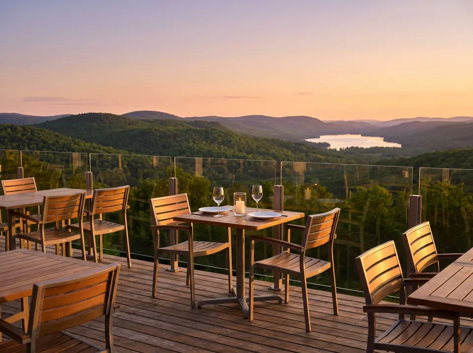 Une terrasse en bois vide à flanc de montagne, une table pour deux avec bougie et verres, et la vue sur des collines boisées et un lac au coucher du soleil.